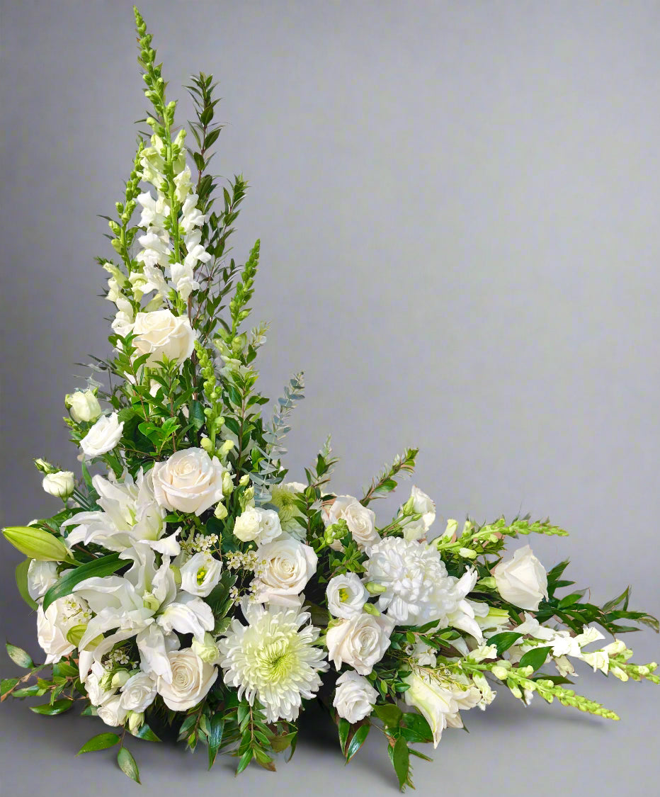 A white floral arrangement featuring a variety of flowers such as roses and lilies, designed in a L-shape for a funeral service.