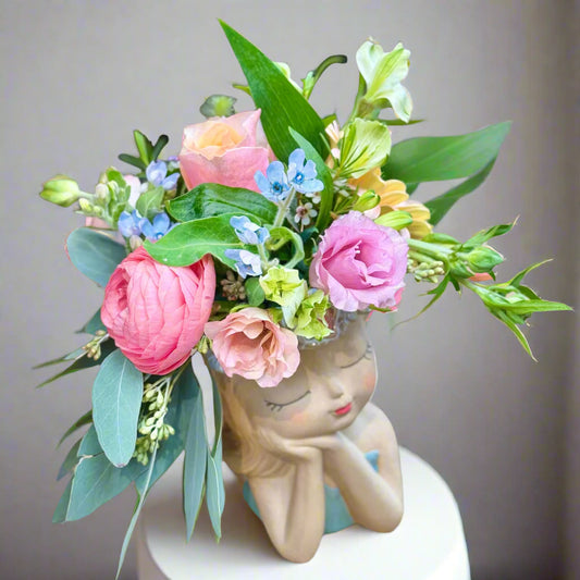 A floral arrangement in the shape of a stylized female figure with a pot as her lap, adorned with various flowers such as pink roses, blue tweedya, and green leaves.