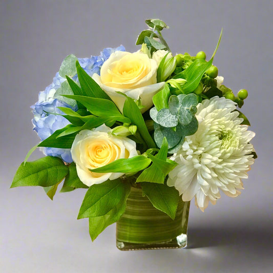 A floral arrangement featuring blue, yellow, and white flowers in a clear glass vase.